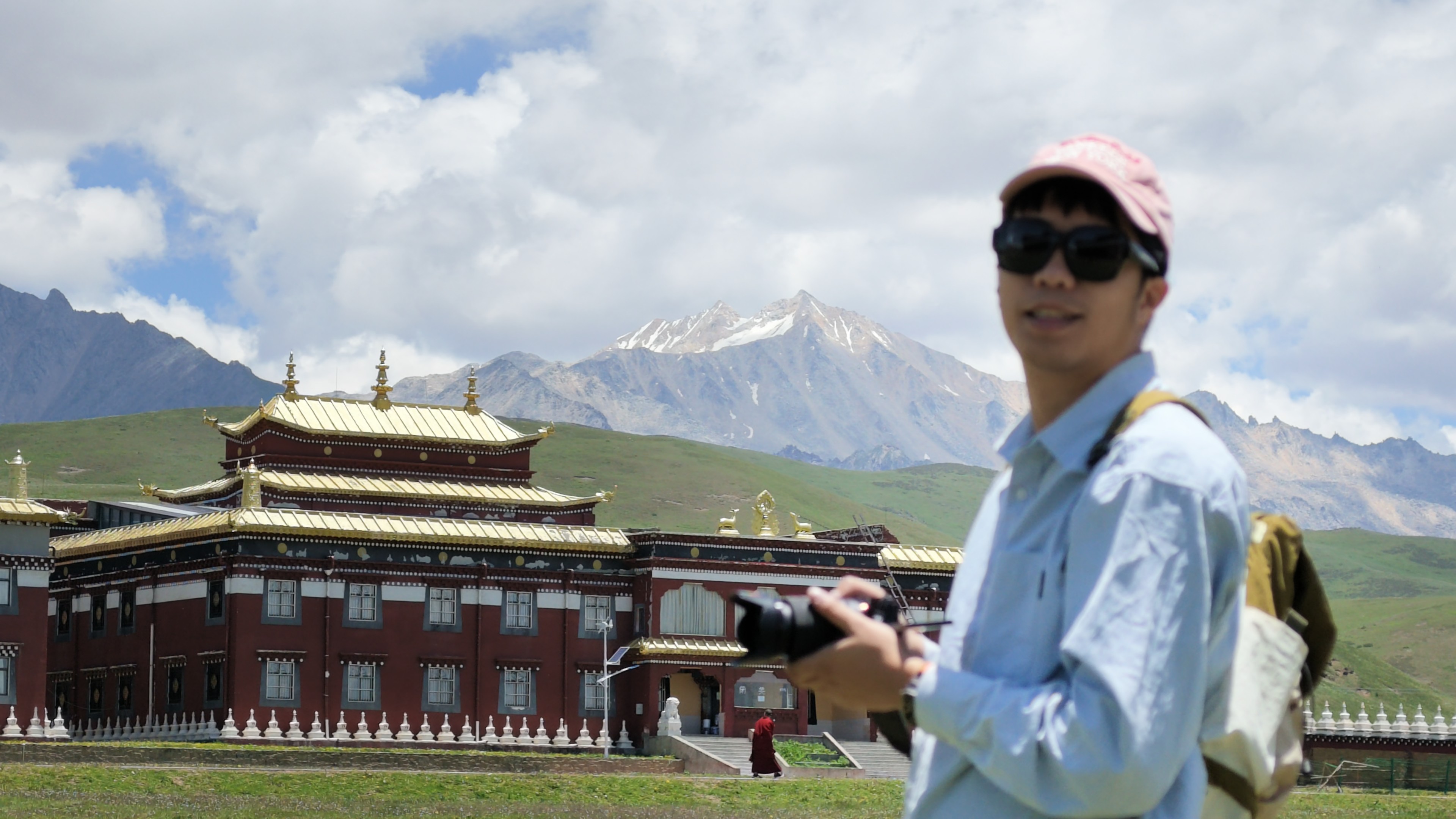 Shenghuan holding a camera outdoors, with snow-capped mountains and a temple in the background